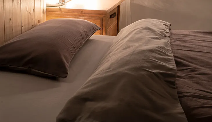 Foto: Rötliches Sonnenlicht scheint in ein dunkles Schlafzimmer auf ein mit Winterbettwäsche aus Flanell bezogenes Bett.
