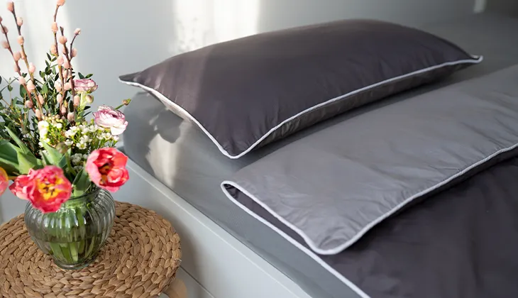 Foto: Ein gemachtes Bett bezogen mit Sommer-Bettwäsche aus Satin, auf dem Nachttisch ein bunter Blumenstrauß.