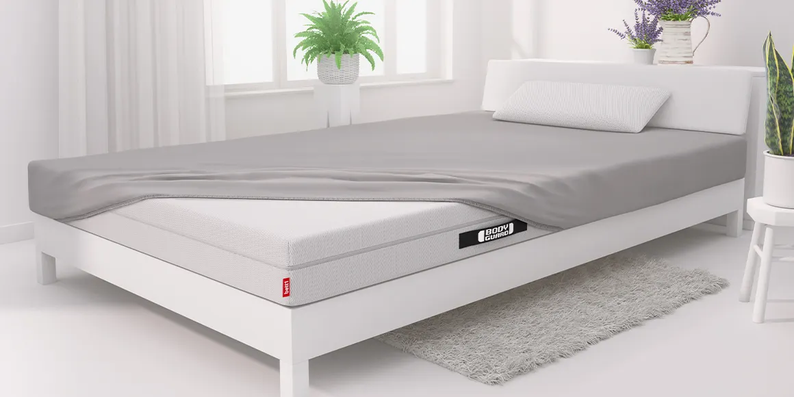 Foto: In einem hellen weißen Schlafzimmer steht ein großes weißes Bett mit BODYGUARD Matratze, die nur an drei Ecken mit einem Spannbettlaken bezogen ist.