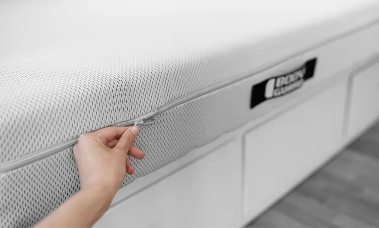 Ręka rozpinająca pokrowiec materaca z napisem BODYGUARD, który leży na łóżku.