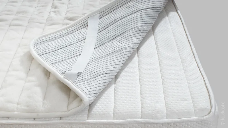 Foto: Auf einer Matratze liegt ein wasserdichter Matratzentopper, eine Ecke ist zurückgeklappt und gibt den Blick auf die Befestigungslasche und die wasserdichte Beschichtung frei.