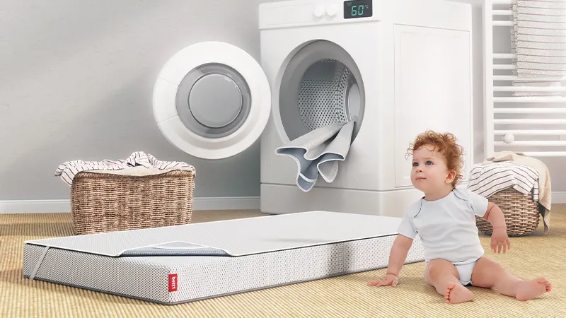 Dziecko siedzące na podłodze obok materaca. Za nim pralka oraz kosz na bieliznę.