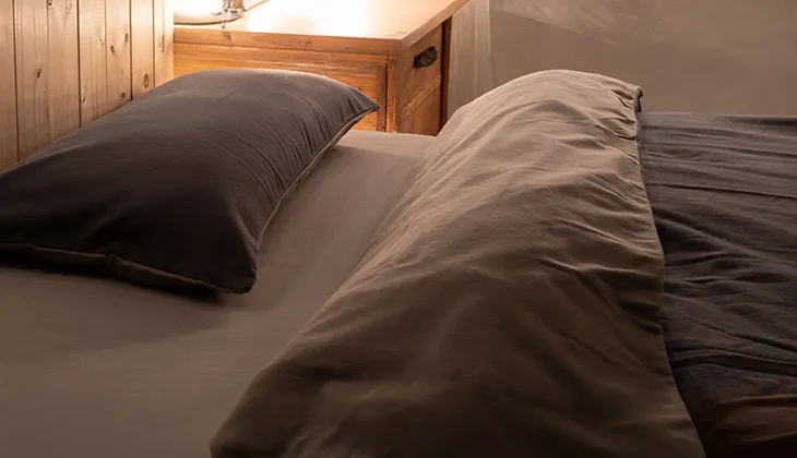 Foto: Rötliches Sonnenlicht scheint in ein dunkles Schlafzimmer auf ein mit Winterbettwäsche aus Flanell bezogenes Bett.