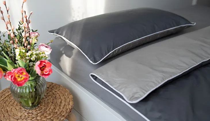 Foto: Ein gemachtes Bett bezogen mit Sommer-Bettwäsche aus Satin, auf dem Nachttisch ein bunter Blumenstrauß.