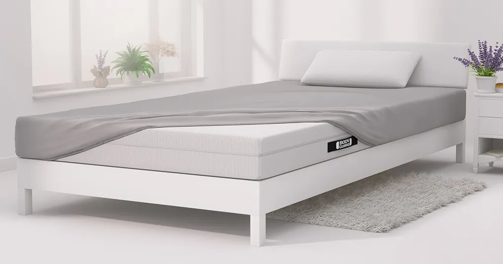 In einem hellen Schlafzimmer steht ein Bett mit BODYGUARD Matratze darauf, die bezogen ist mit BODYGUARD Jersey-Spannbettlaken in Silber.