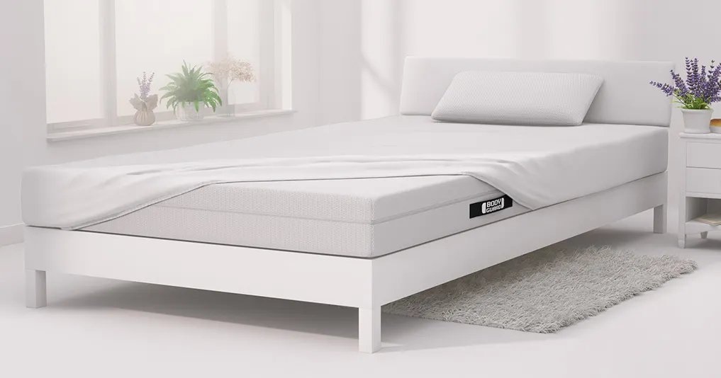 In einem hellen Schlafzimmer steht ein Bett mit BODYGUARD Matratze darauf, die bezogen ist mit BODYGUARD Jersey-Spannbettlaken in Weiß.