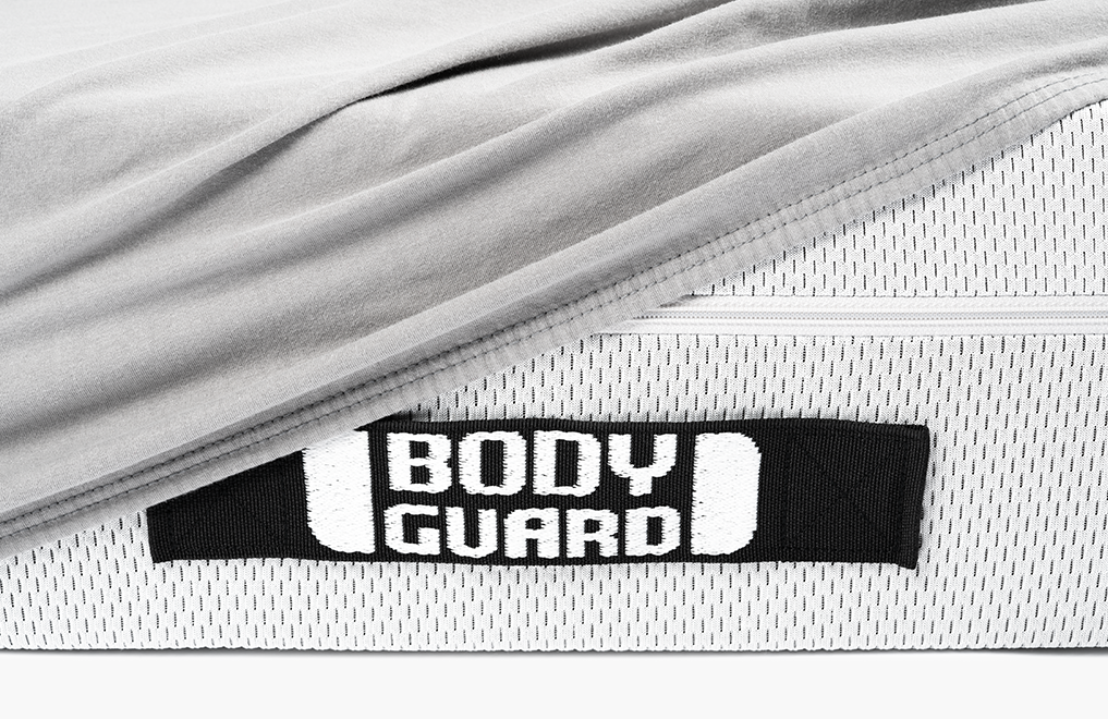 Fragment materaca BODYGUARD z widocznym logo oraz częściowo naciągniętym szarym prześcieradłem z gumką.